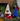 Small dog sitting among festive Christmas decorations including a tree-shaped pet bed and colorful felt trees.