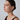 A close-up side profile of a woman with dark hair pulled back, wearing a delicate gold hoop earring with small clear stones, against a light gray studio background.