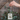 A hand holds a rectangular metal dog tag with a brown hanging loop and white bead. The tag has a pale blue-gray background with cute dog illustrations and the words 'DOG MOM' in pink.