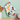 A pair of light blue oven mitts with a colorful Christmas-themed print of cats, hanging on a wall. The background is a soft mint green wall with wooden kitchen utensils on the left and a white enamel pot on the right.