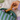 A hand with lavender nail polish holds a small glossy dachshund-shaped cosmetic container above an open green and light blue striped zip pouch on a white fabric surface.